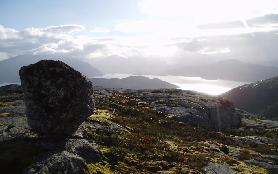 A large boulder in the foreground overlooks a sunlit fjord with mountains in the distance under a cloudy sky.
